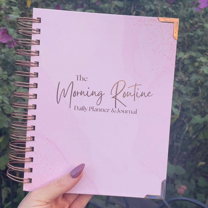 May include: A pink daily planner and journal with a gold spiral binding and the text "The Morning Routine Daily Planner & Journal" on the front cover.