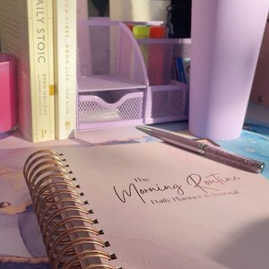 May include: A pink and white daily planner and journal with gold spiral binding. The journal has the text "The Morning Routine Daily Planner & Journal" on the cover. The journal is on a table with a purple cup, a pen, and two books.