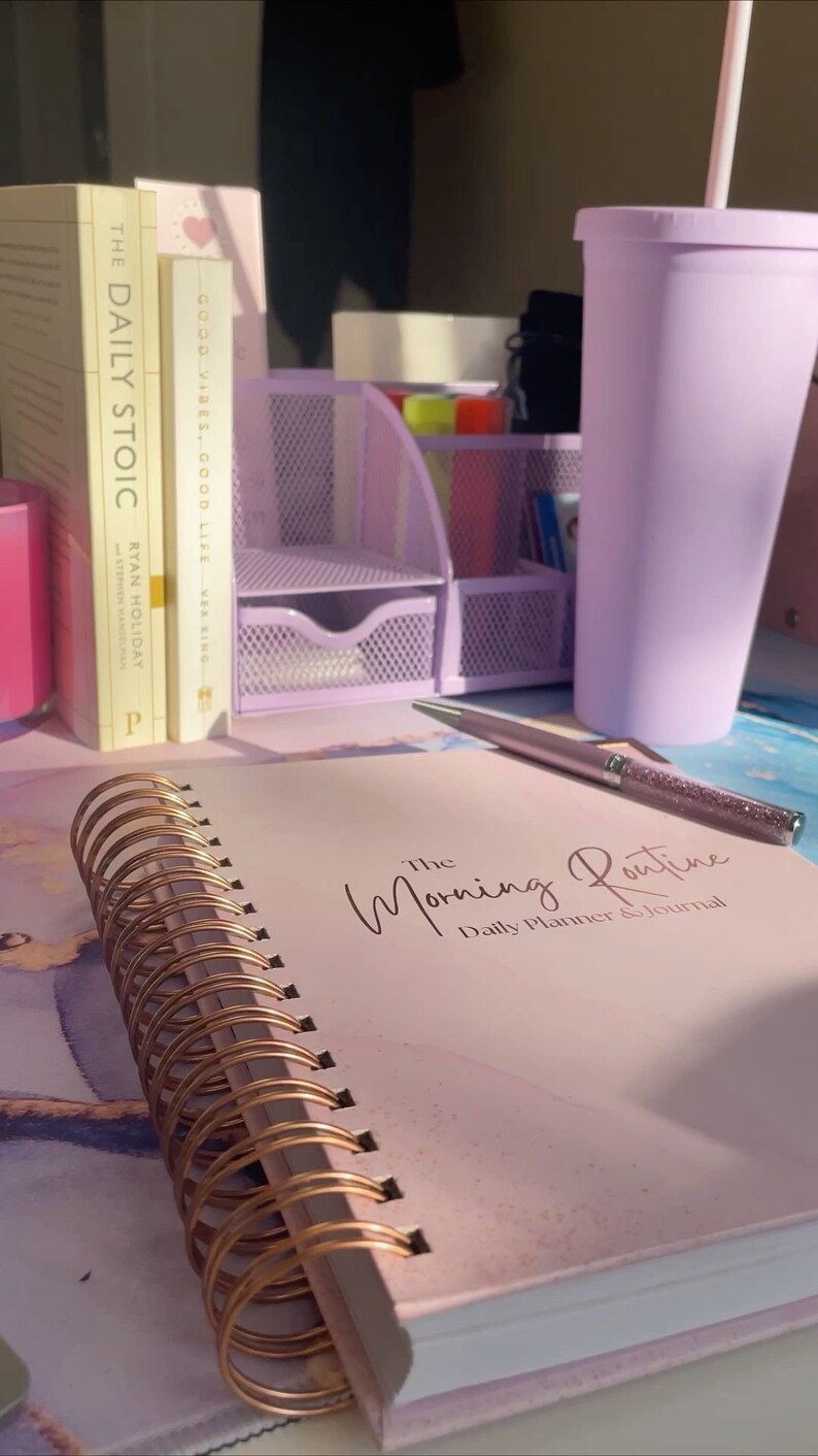 May include: A pink and white daily planner and journal with gold spiral binding. The journal has the text "The Morning Routine Daily Planner & Journal" on the cover. The journal is on a table with a purple cup, a pen, and two books.