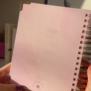 May include: A pink spiral bound notebook with a white marble design and gold accents. The notebook has a small heart with the words "supplied with" around it.