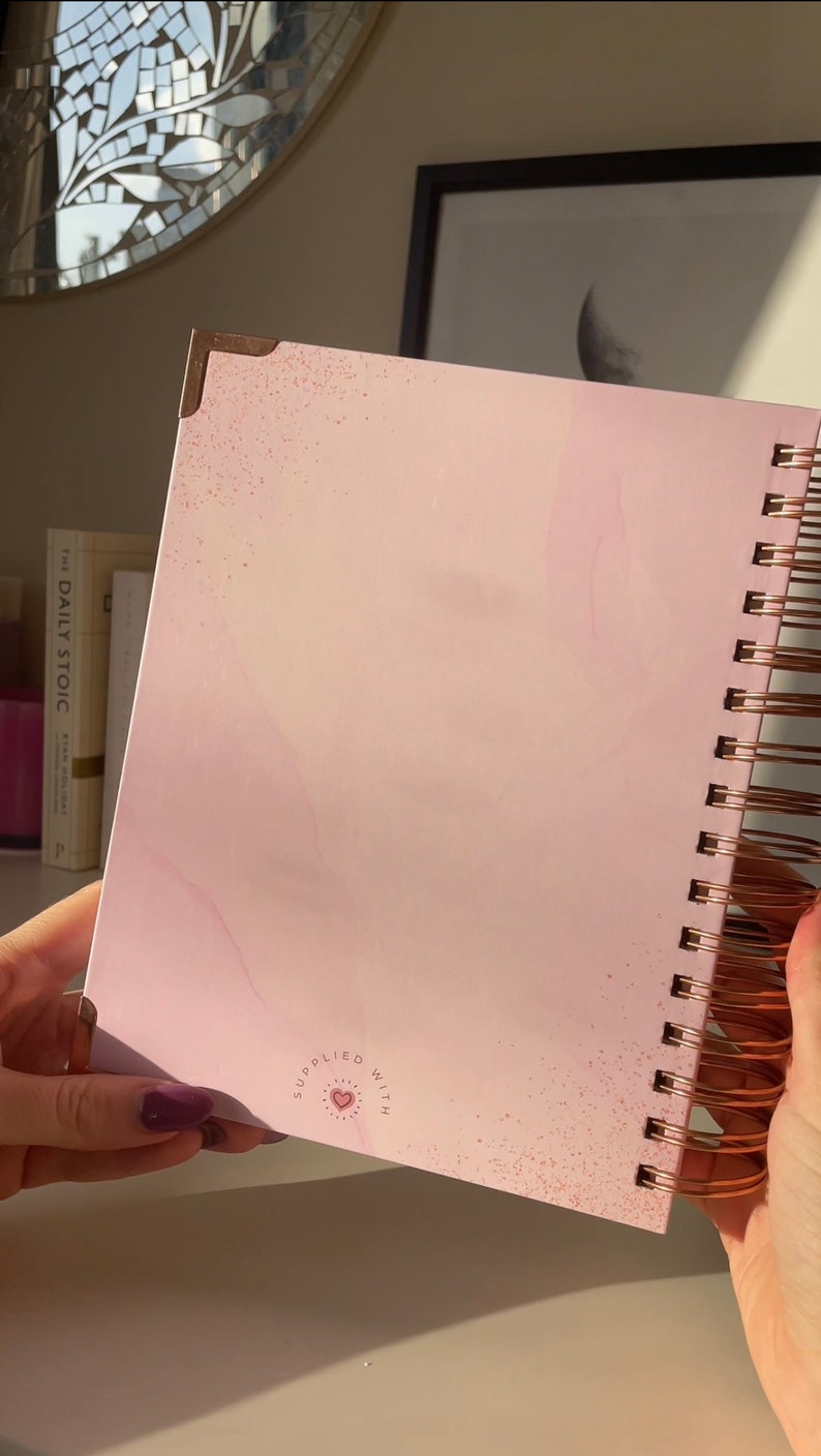 May include: A pink spiral bound notebook with a white marble design and gold accents. The notebook has a small heart with the words "supplied with" around it.