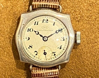 Può includere: Un orologio da polso in argento con quadrante bianco e lancette nere. L'orologio ha un cinturino in pelle marrone e il testo "IWC Schaffhausen" sul quadrante.