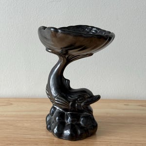 May include: A dark grey ceramic dolphin ornament with a bowl-shaped top. The dolphin is sculpted with detailed fins and a smiling face, resting on a textured base. The bowl is scalloped around the rim.
