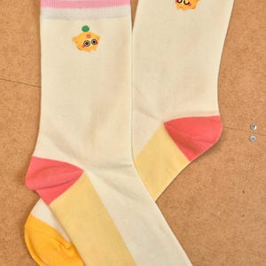 K&ouml;nnte beinhalten: Ein Paar cremefarbene Socken mit Cupcake-Design. Die Socken haben rosa und gelbe Akzente an Zehen und Ferse sowie einen rosa Streifen oben. Ein Cartoon-Cupcake ist oben aufgestickt.
