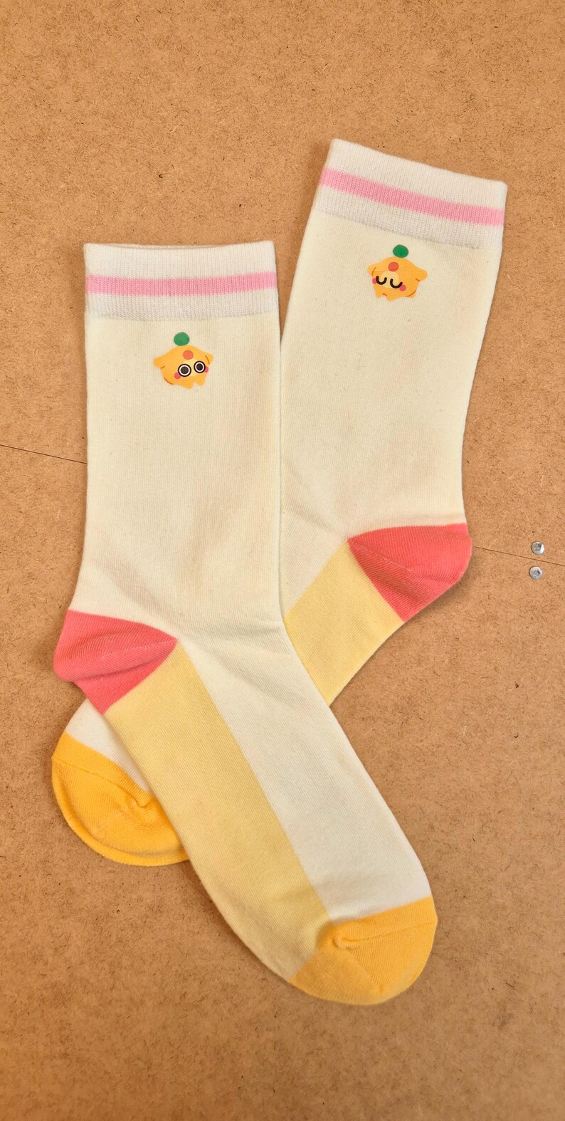K&ouml;nnte beinhalten: Ein Paar cremefarbene Socken mit Cupcake-Design. Die Socken haben rosa und gelbe Akzente an Zehen und Ferse sowie einen rosa Streifen oben. Ein Cartoon-Cupcake ist oben aufgestickt.