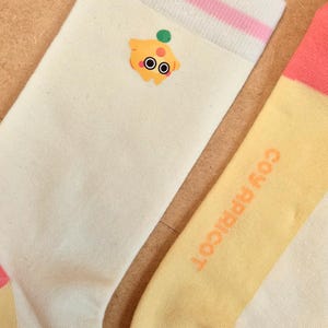 K&ouml;nnte beinhalten: Ein Paar Socken mit einem Cartoon-Aprikosen-Design. Eine Socke ist cremefarben mit einem rosa und wei&szlig;en gestreiften B&uuml;ndchen und einer Cartoon-Aprikose. Die andere Socke ist gelb mit dem Text "COY APRICOT" in Orange.