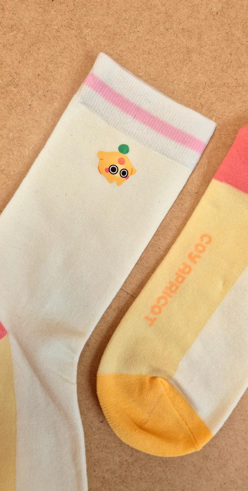 K&ouml;nnte beinhalten: Ein Paar Socken mit einem Cartoon-Aprikosen-Design. Eine Socke ist cremefarben mit einem rosa und wei&szlig;en gestreiften B&uuml;ndchen und einer Cartoon-Aprikose. Die andere Socke ist gelb mit dem Text "COY APRICOT" in Orange.