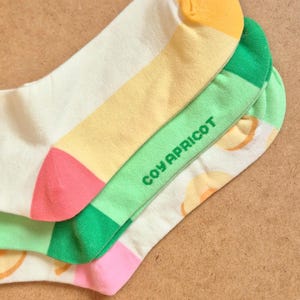 K&ouml;nnte beinhalten: Ein Paar bunte Socken mit Fruchtthema. Die Socken haben eine wei&szlig;e Basis mit gelben, gr&uuml;nen und rosa Akzenten. Der Text "COY APRICOT" ist auf dem gr&uuml;nen Abschnitt aufgedruckt. Die Socken sind aus weichem Material.