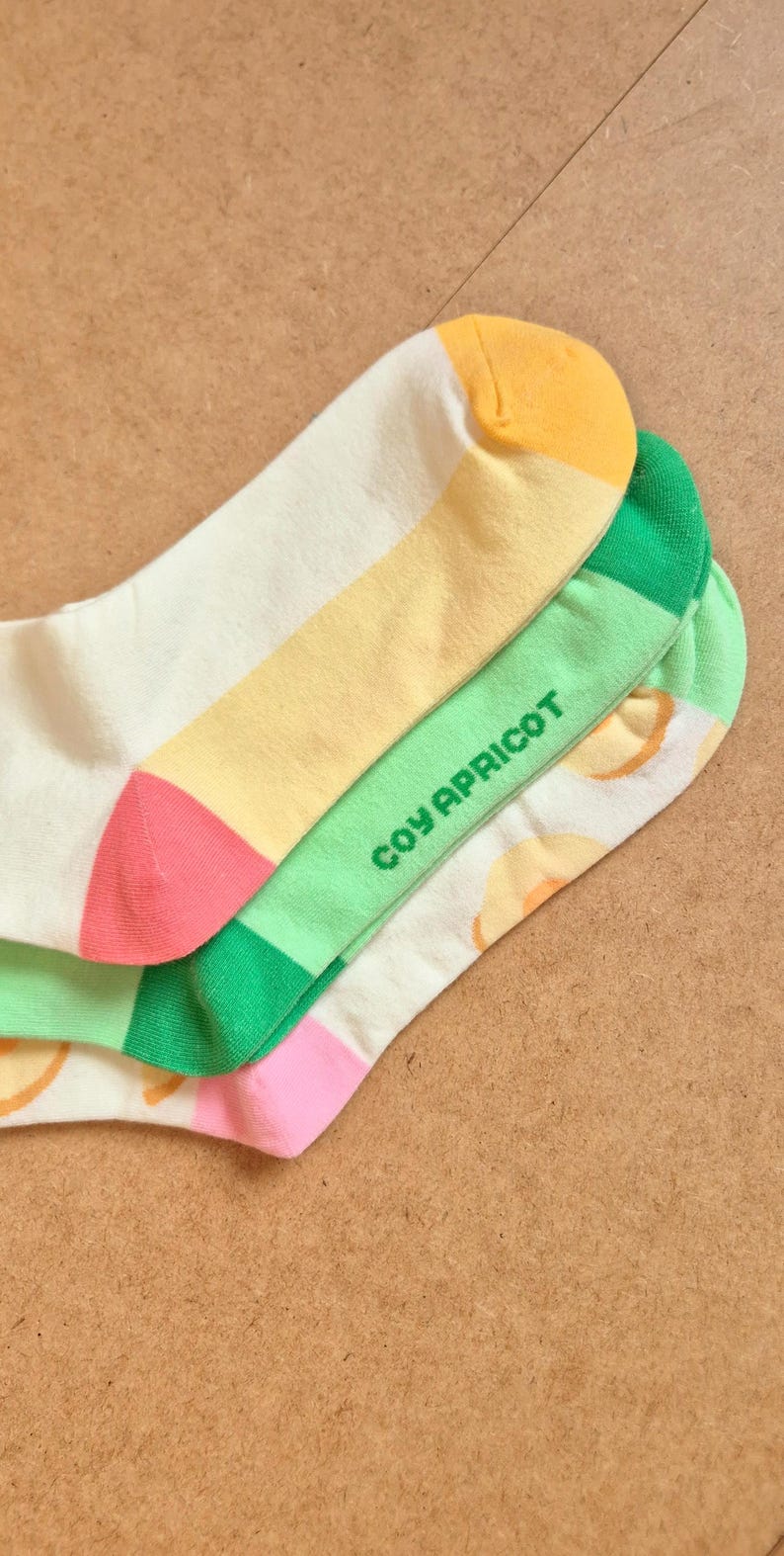 K&ouml;nnte beinhalten: Ein Paar bunte Socken mit Fruchtthema. Die Socken haben eine wei&szlig;e Basis mit gelben, gr&uuml;nen und rosa Akzenten. Der Text "COY APRICOT" ist auf dem gr&uuml;nen Abschnitt aufgedruckt. Die Socken sind aus weichem Material.