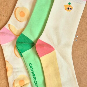 K&ouml;nnte beinhalten: Drei Paar bunte Socken mit Obst- und Cartoon-Designs. Ein Paar hat Pfirsichscheiben, ein anderes einen Cartoon-Charakter und das dritte den Text "COY APRICOT". Die Socken haben rosa, gr&uuml;ne und gelbe Akzente.