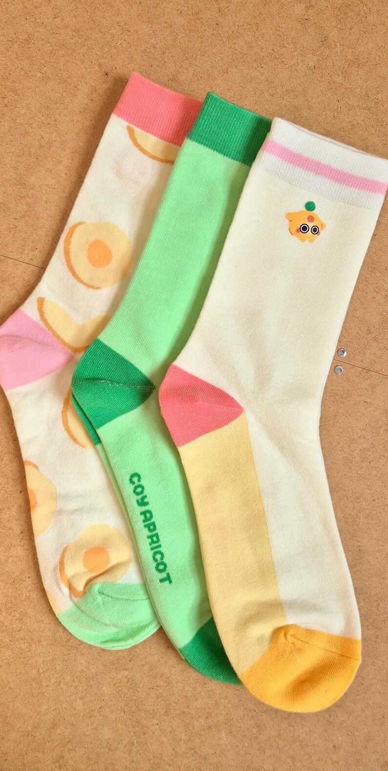 K&ouml;nnte beinhalten: Drei Paar bunte Socken mit Obst- und Cartoon-Designs. Ein Paar hat Pfirsichscheiben, ein anderes einen Cartoon-Charakter und das dritte den Text "COY APRICOT". Die Socken haben rosa, gr&uuml;ne und gelbe Akzente.
