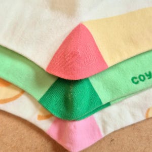 K&ouml;nnte beinhalten: Nahaufnahme von bunten Socken. Die Socken sind wei&szlig; mit rosa, gelben und gr&uuml;nen Akzenten. Das Wort "coy" ist auf einer der Socken zu sehen. Die Socken sind gefaltet und gestapelt.