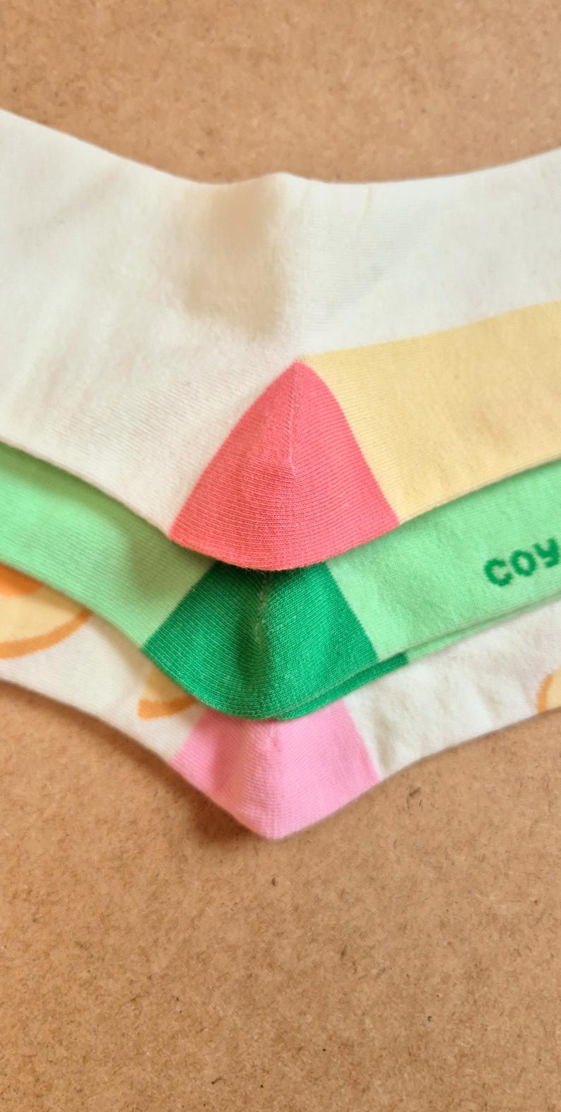 K&ouml;nnte beinhalten: Nahaufnahme von bunten Socken. Die Socken sind wei&szlig; mit rosa, gelben und gr&uuml;nen Akzenten. Das Wort "coy" ist auf einer der Socken zu sehen. Die Socken sind gefaltet und gestapelt.