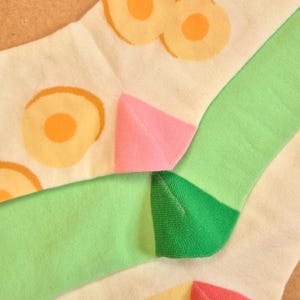 K&ouml;nnte beinhalten: Nahaufnahme von bunten Socken mit Fruchtmuster. Die Socken haben eine wei&szlig;e Basis mit orangefarbenen und gelben Fruchtmustern, gr&uuml;nen und rosa Akzenten und dem Text "COY APRICOT" in Orange.