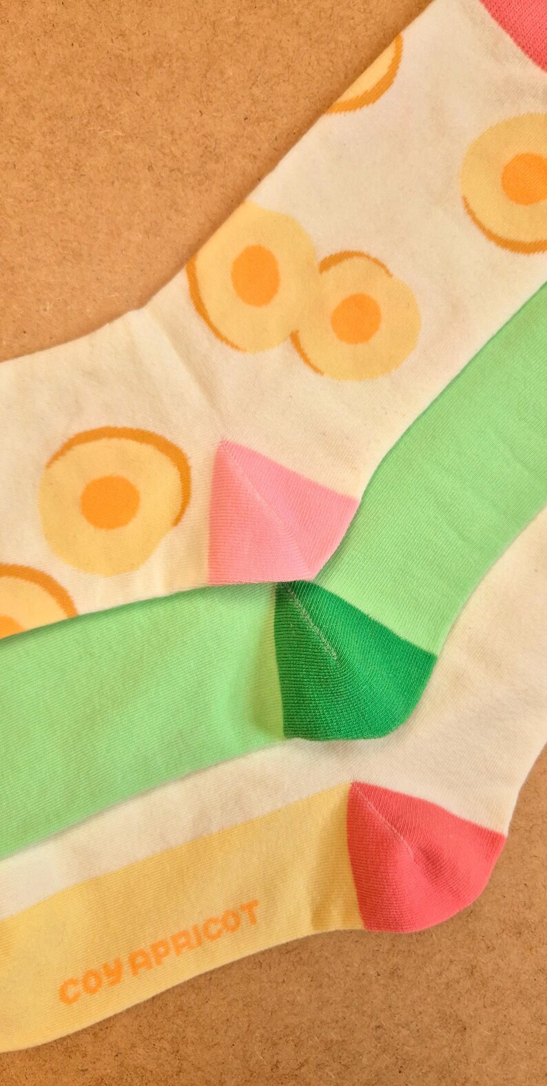 K&ouml;nnte beinhalten: Nahaufnahme von bunten Socken mit Fruchtmuster. Die Socken haben eine wei&szlig;e Basis mit orangefarbenen und gelben Fruchtmustern, gr&uuml;nen und rosa Akzenten und dem Text "COY APRICOT" in Orange.