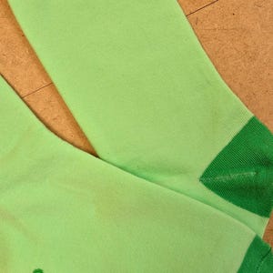 K&ouml;nnte beinhalten: Ein Paar hellgr&uuml;ne Socken mit dunkelgr&uuml;nen Akzenten an der Oberseite und den Zehen. Der Text "COY APRICOT" ist gr&uuml;n auf der Seite einer Socke aufgedruckt. Die Socken sind aus weichem Rippmaterial.