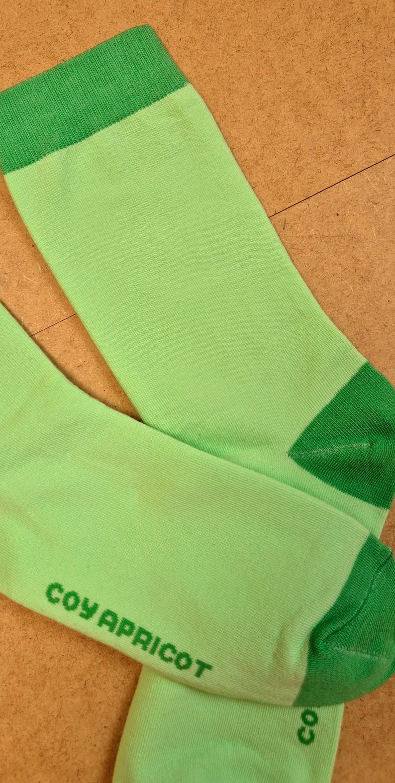 K&ouml;nnte beinhalten: Ein Paar hellgr&uuml;ne Socken mit dunkelgr&uuml;nen Akzenten an der Oberseite und den Zehen. Der Text "COY APRICOT" ist gr&uuml;n auf der Seite einer Socke aufgedruckt. Die Socken sind aus weichem Rippmaterial.