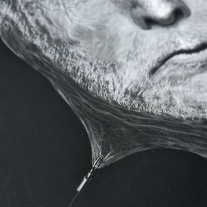 May include: A black and white drawing of a man's face, with a white string attached to the bottom of the drawing. The string is tied to a small metal hook.