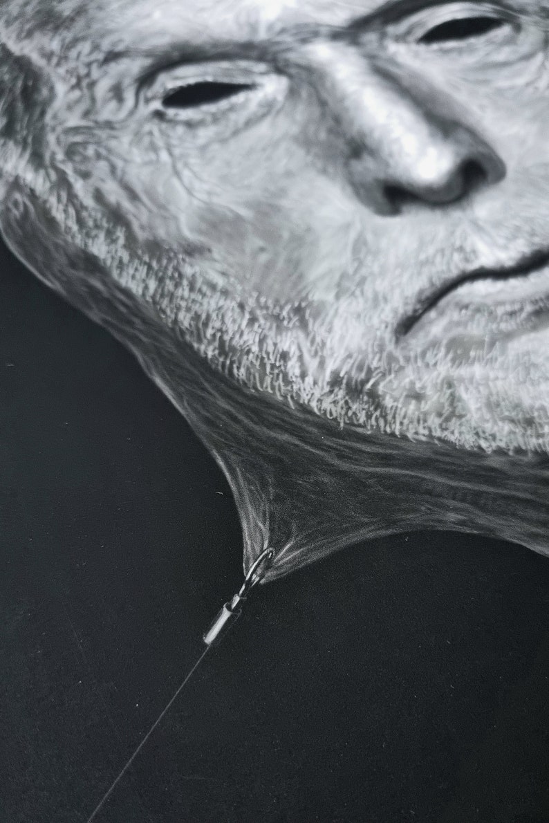 May include: A black and white drawing of a man's face, with a white string attached to the bottom of the drawing. The string is tied to a small metal hook.