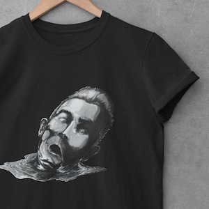 Op de afbeelding: Zwart T-shirt met een witte grafische print van een mannengezicht met een verontruste uitdrukking. Het gezicht van de man is gedeeltelijk ondergedompeld in water.