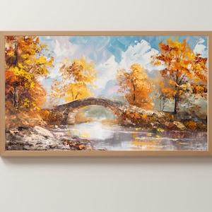 May include: Oil painting of a stone bridge over a calm river, framed in a light wood. The artwork depicts an autumn landscape with trees in shades of orange and yellow, under a cloudy blue sky.