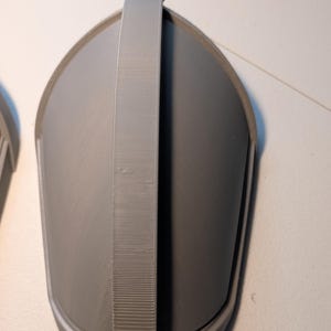 May include: A gray, 3D-printed, shield-shaped armor component featuring a smooth, curved surface and a prominent, textured vertical strap running down the center. The object has a rounded top and tapers toward the bottom, with a raised border edge.