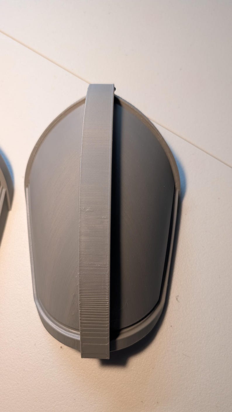 May include: A gray, 3D-printed, shield-shaped armor component featuring a smooth, curved surface and a prominent, textured vertical strap running down the center. The object has a rounded top and tapers toward the bottom, with a raised border edge.