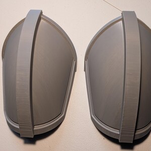 May include: A pair of matching gray shoulder pauldrons featuring a smooth, curved dome shape with a prominent central vertical ridge running from top to bottom. These rigid, molded components have a clean, matte finish and a symmetrical design.