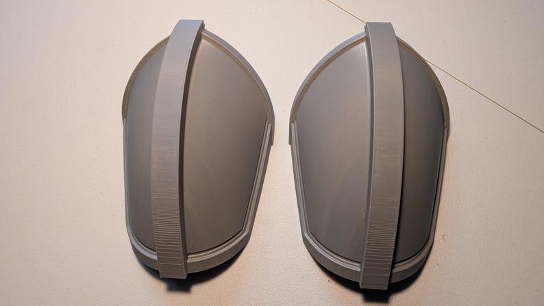 May include: A pair of matching gray shoulder pauldrons featuring a smooth, curved dome shape with a prominent central vertical ridge running from top to bottom. These rigid, molded components have a clean, matte finish and a symmetrical design.