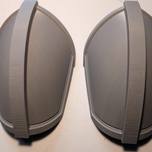 May include: A pair of matching gray shoulder pauldrons featuring a smooth, curved surface and a prominent central ridge that runs vertically along the length of each piece. The armor components have a clean, matte finish and a structured design.