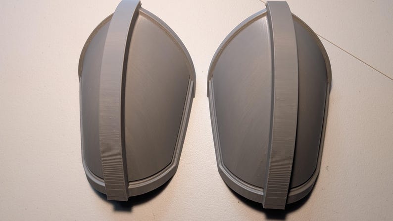 May include: A pair of matching gray shoulder pauldrons featuring a smooth, curved surface and a prominent central ridge that runs vertically along the length of each piece. The armor components have a clean, matte finish and a structured design.