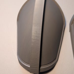 May include: A gray, teardrop-shaped protective guard featuring a smooth, curved surface and a central vertical strap running from top to bottom. The object has a matte finish and a distinct raised border along its outer edge, showing a 3D printed texture.
