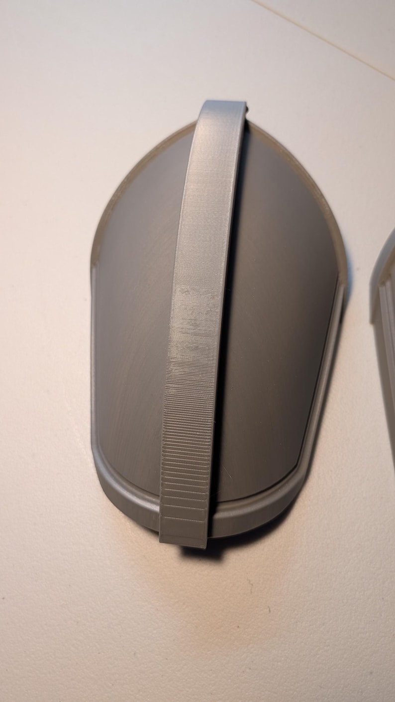 May include: A gray, teardrop-shaped protective guard featuring a smooth, curved surface and a central vertical strap running from top to bottom. The object has a matte finish and a distinct raised border along its outer edge, showing a 3D printed texture.