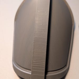 May include: A matte grey, 3D-printed, dome-shaped object featuring a smooth, curved exterior surface and a prominent, vertical, textured strap running across the center from top to bottom, with a defined base rim and a solid, uniform construction.