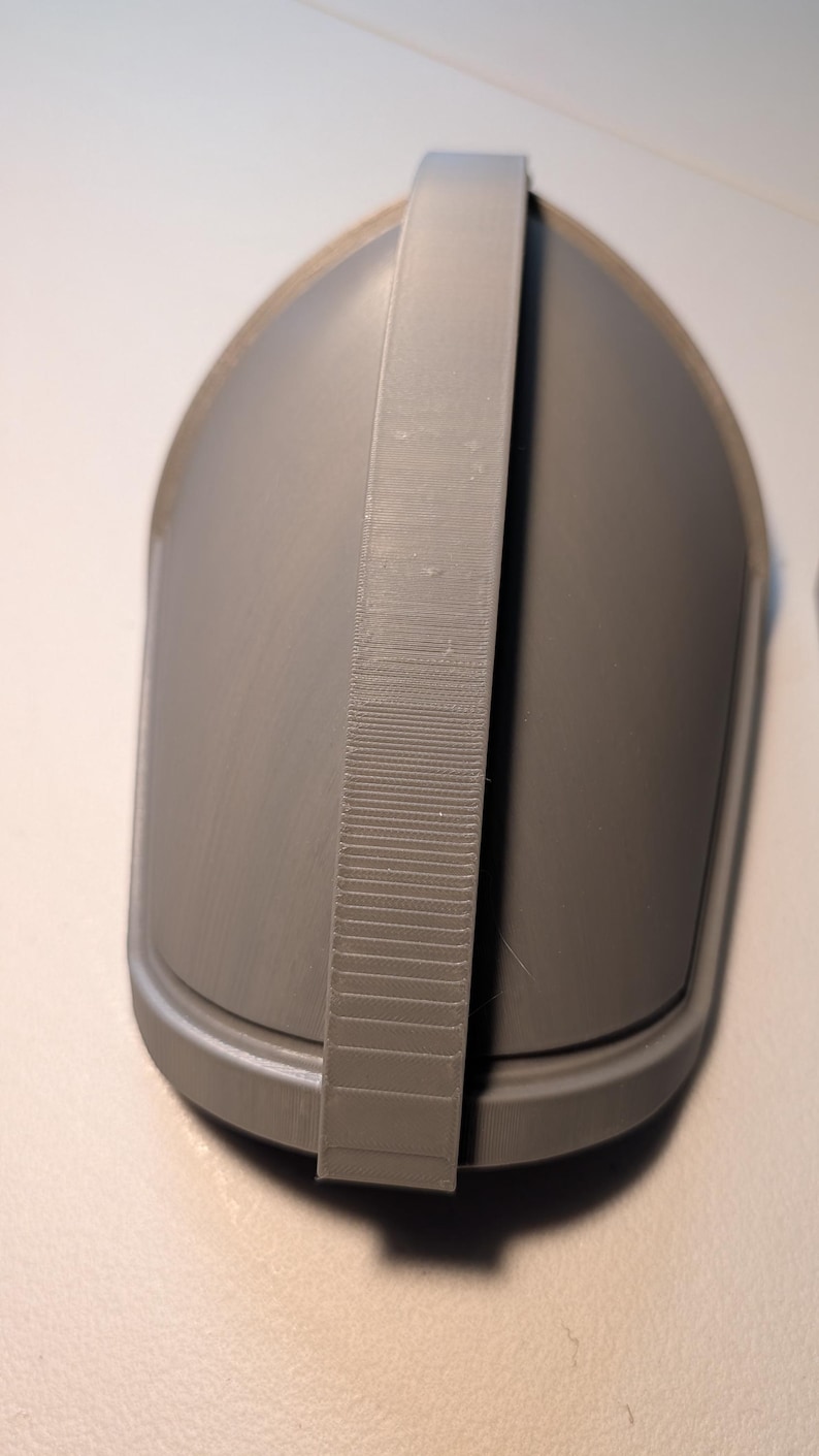 May include: A matte grey, 3D-printed, dome-shaped object featuring a smooth, curved exterior surface and a prominent, vertical, textured strap running across the center from top to bottom, with a defined base rim and a solid, uniform construction.