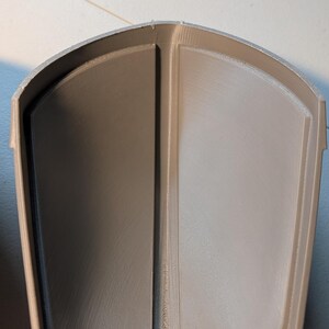 May include: A light gray, 3D-printed, curved, shield-like component with a smooth, matte finish. The object features a central vertical ridge that divides the interior into two symmetrical, concave sections, with a raised outer border.