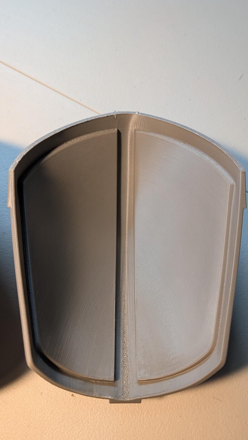 May include: A light gray, 3D-printed, curved, shield-like component with a smooth, matte finish. The object features a central vertical ridge that divides the interior into two symmetrical, concave sections, with a raised outer border.