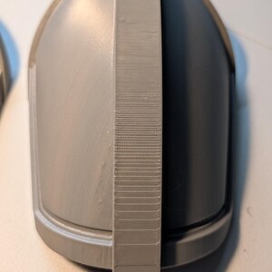 May include: A gray, dome-shaped object with a smooth, curved surface and a central, vertical ridge featuring a textured, layered finish. The piece has a rounded base and a solid, uniform gray color throughout its entire structure.
