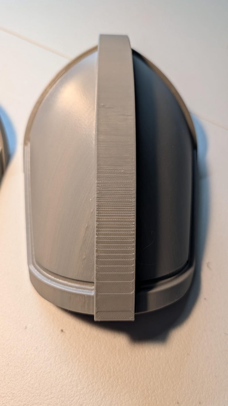 May include: A gray, dome-shaped object with a smooth, curved surface and a central, vertical ridge featuring a textured, layered finish. The piece has a rounded base and a solid, uniform gray color throughout its entire structure.