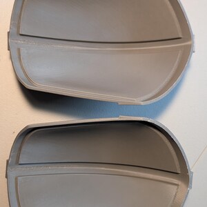 May include: Two identical gray, curved, semi-rigid components with a smooth, matte finish. Each piece features a raised central horizontal ridge and a defined, slightly elevated border along the perimeter, creating a concave, shell-like structure.