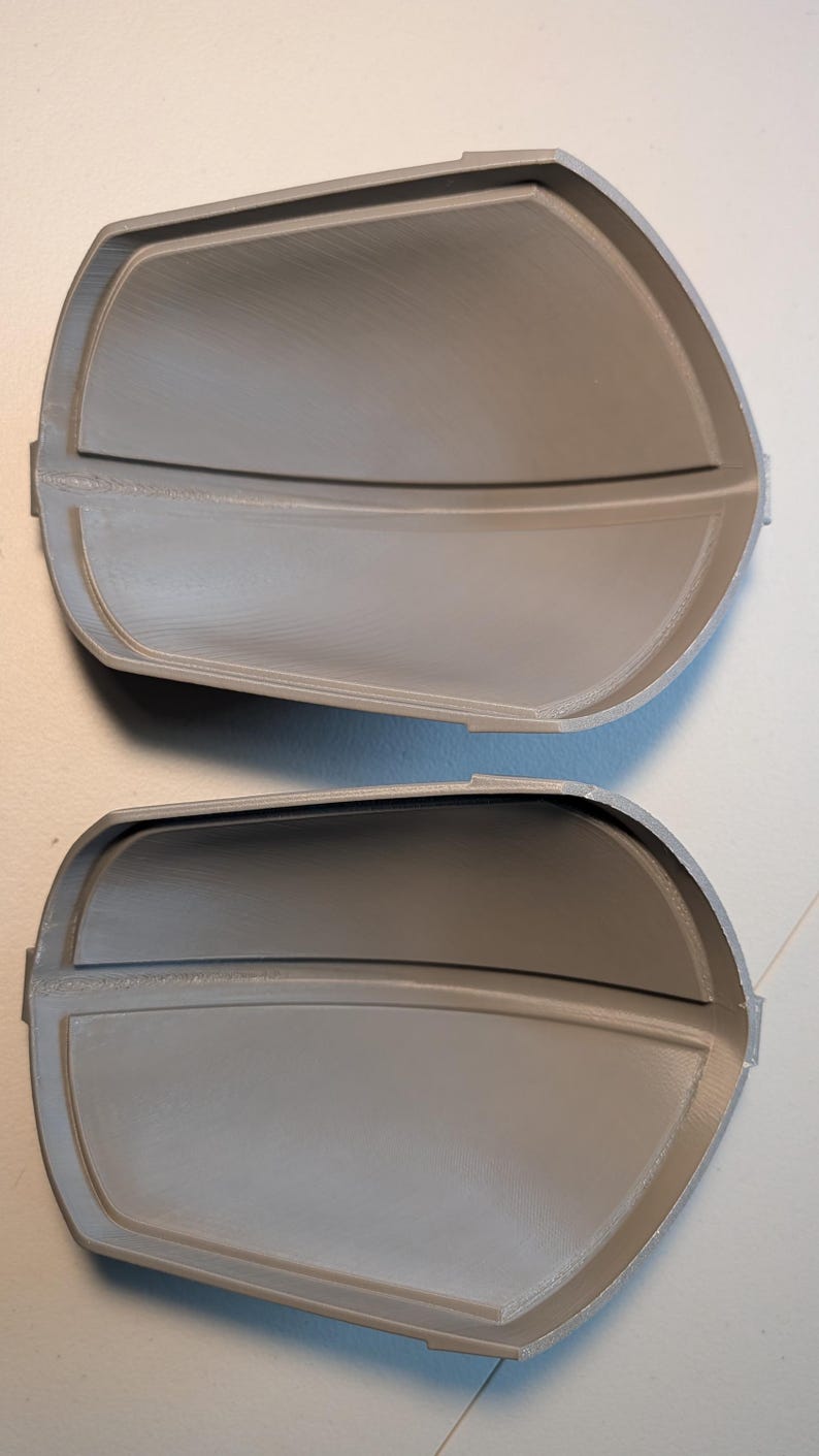 May include: Two identical gray, curved, semi-rigid components with a smooth, matte finish. Each piece features a raised central horizontal ridge and a defined, slightly elevated border along the perimeter, creating a concave, shell-like structure.