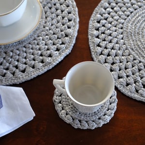 Puede incluir: Un conjunto de manteles individuales, posavasos y un servilletero plateados de ganchillo. Una taza de té blanca se encuentra sobre un posavasos y otra sobre un mantel individual. También hay una servilleta con un diseño de ancla azul.