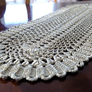 May include: A silver-colored, oval-shaped table runner. The runner is made of a crocheted material with a detailed, openwork design. The edges have a scalloped trim. The runner is placed on a dark brown wooden table.