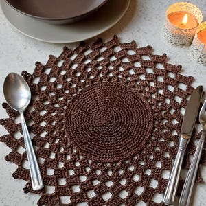 Pode incluir: Um tapete de crochê castanho com um design circular. O tapete é feito com um padrão delicado e aberto e tem uma textura lisa e uniforme.