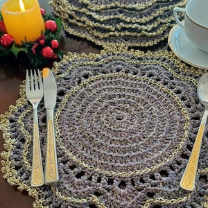 May include: A set of handmade crochet placemats and coasters in shades of gray with a gold trim. The table setting includes silverware, a teacup, and a lit candle with festive decorations. The placemats are circular.