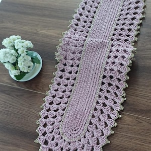 Może przedstawiać: Długi, owalny bieżnik stołowy wykonany na szydełku w jasnoróżowym kolorze z złotą lamówką. Bieżnik wykonany jest z delikatnego, ażurowego wzoru.