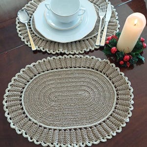 May include: Oval, crocheted placemats in a neutral beige color with a scalloped edge. The placemats are set on a dark brown table, with white plates, a cup, silverware, and a lit candle.