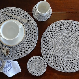 Può includere: Un set di tovagliette e sottobicchieri grigi all'uncinetto, con una tazza da tè e un piattino bianchi, un cucchiaio e una forchetta. Un tovagliolo bianco con un disegno di ancora blu è tenuto da un anello per tovaglioli all'uncinetto. Gli oggetti sono su una superficie di legno scuro.