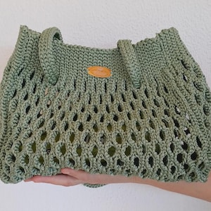 Op de afbeelding: Een groene gehaakte draagtas met een bruin leren label. De tas heeft een gaasontwerp en is perfect om boodschappen of andere spullen in te dragen.