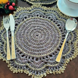 Pode incluir: Um conjunto de tapetes de crochê com acabamento dourado. Os tapetes são cinzentos e têm um design delicado e rendado. Um garfo, uma faca e uma colher estão colocados em um dos tapetes.